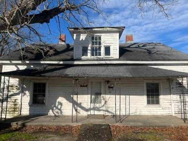 A weathered white single-story house with black shingles and a metal awning, displaying some deterioration, under a clear sky with the shadow of a leafless tree cast upon it.
