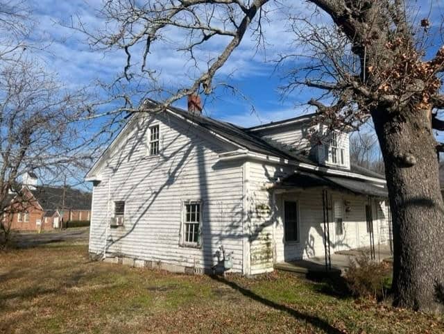 A white, single-story house with weathered siding stands under a bare tree, with shadows cast on its facade under a clear sky.