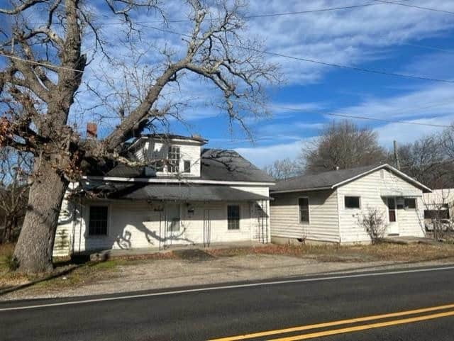 A large tree has fallen onto a white two-story house, causing significant damage to the roof and upper structure, next to a road under a clear sky.