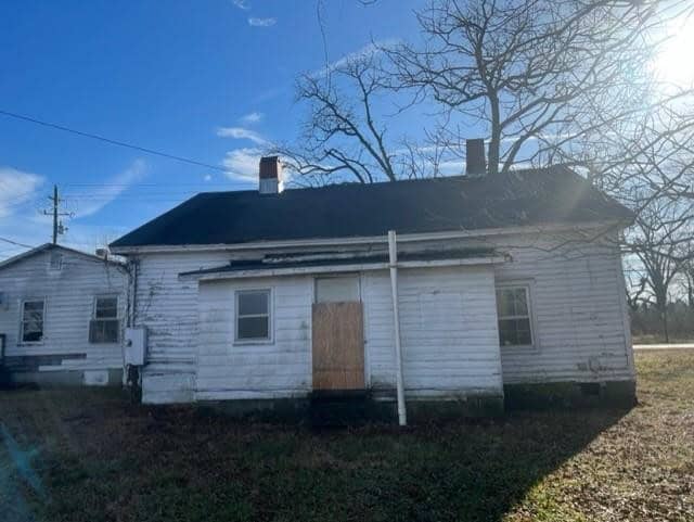A small, white, single-story house in disrepair under a clear sky, with boarded-up windows and a neglected appearance.