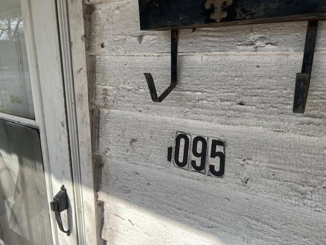 A weathered wall with the numbers 095 mounted on it, next to a white door with a metal handle, and a black hook above the numbers.