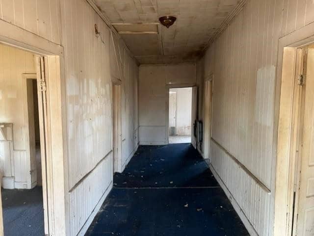A dimly lit, narrow corridor with wooden paneling on the walls and several closed doors leading off to the sides. the floor appears to be covered with a dark carpet, and there are visible stains or discolorations on the walls. the passage seems old and somewhat neglected.