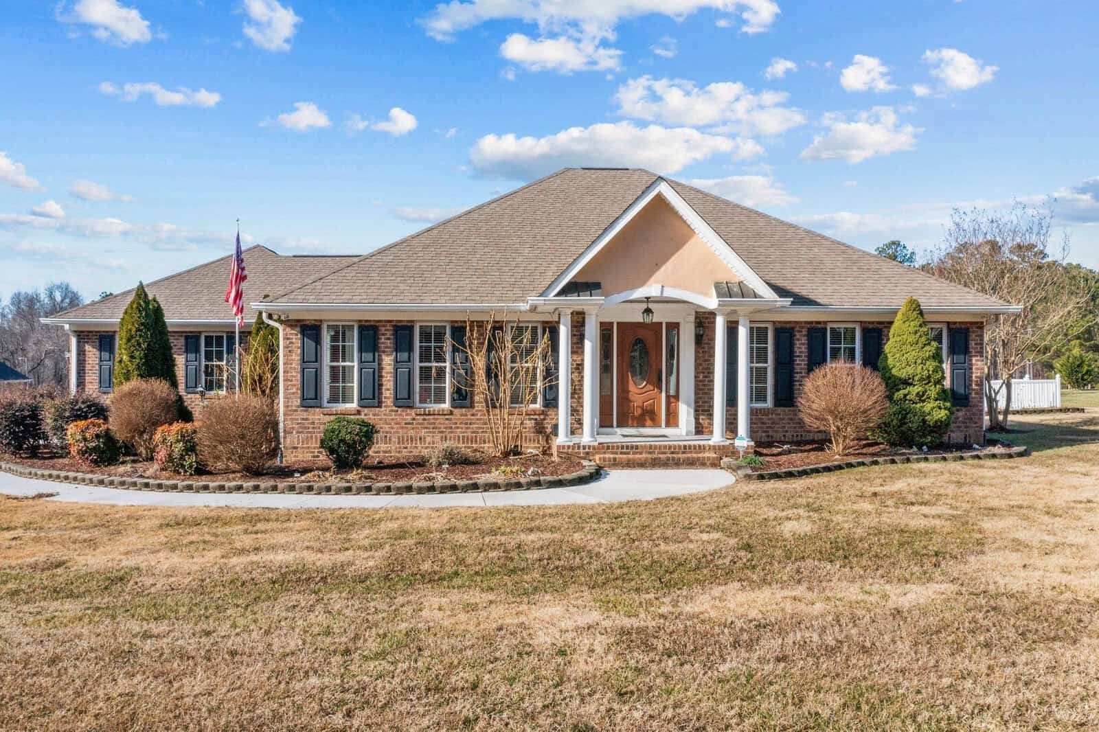 A single-story suburban home at 22 Greta Lane, Henderson NC, with a brown shingle roof, brick facade, and a landscaped yard with a paved pathway leading to an arched front