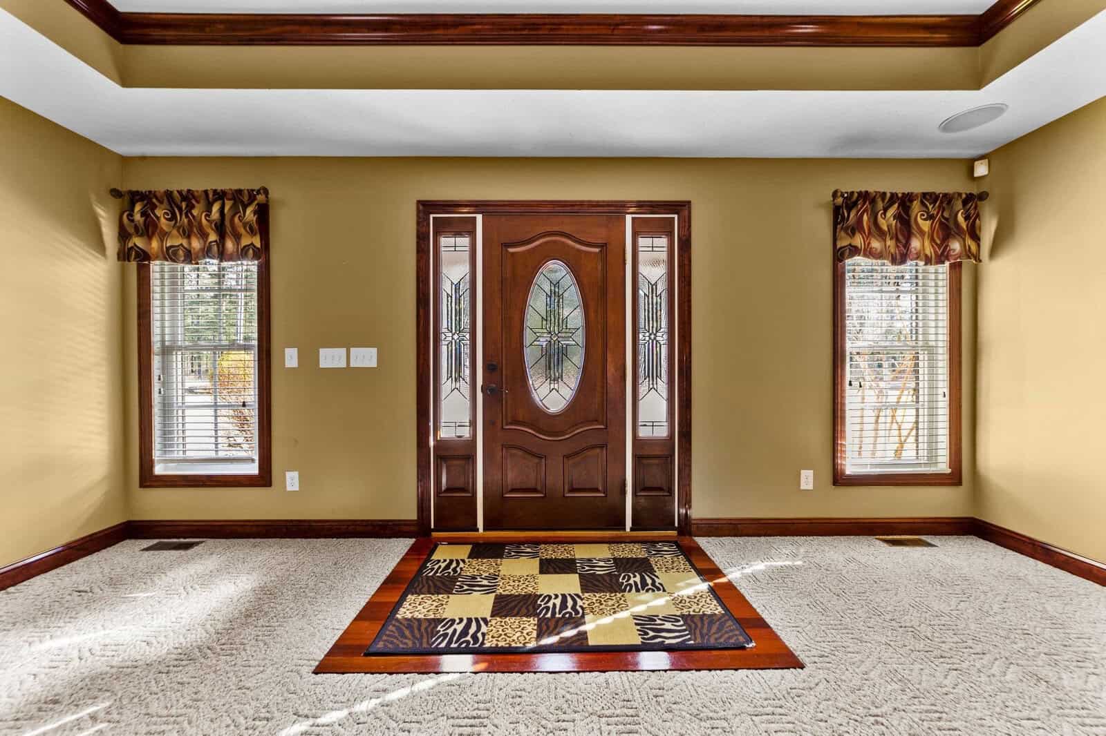 An empty room with a decorative wooden front door featuring stained glass, flanked by two windows with patterned drapes, and a tiled area resembling a chessboard design at the entrance.