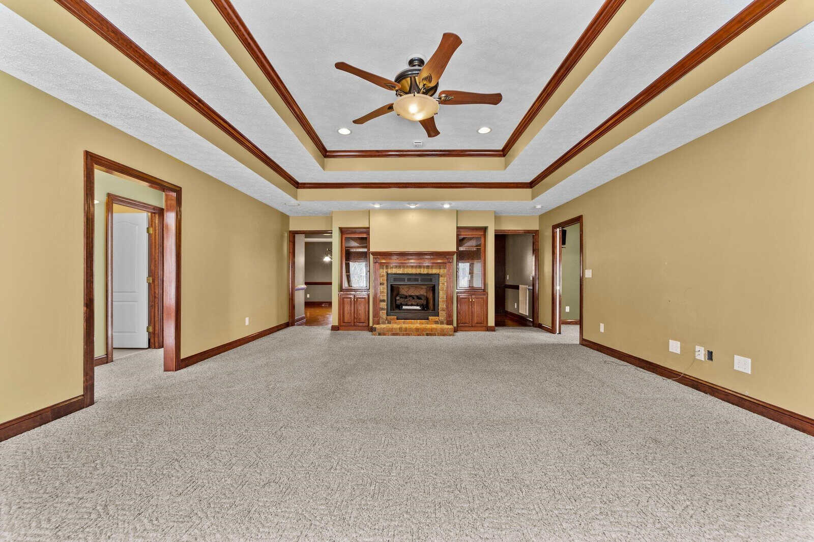 Spacious living room featuring a beige color scheme, carpeted flooring, a ceiling fan, recessed lighting, and a central fireplace with surrounding built-in shelving.