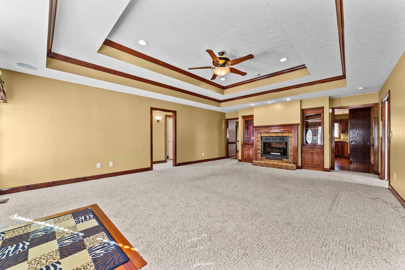 A spacious living room with beige walls and white crown molding, featuring a fireplace with a wood mantle and built-in shelving, under a ceiling with exposed dark wood beams and a ceiling fan.