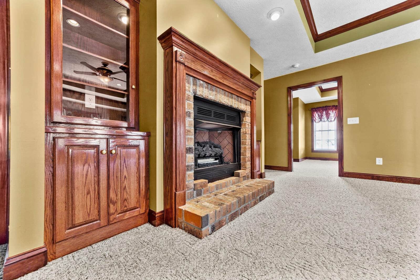 A spacious room featuring a brick fireplace with a wooden mantel, built-in shelving, carpeted flooring, and green walls with white trim, leading to an adjoining room with a purple curtain.