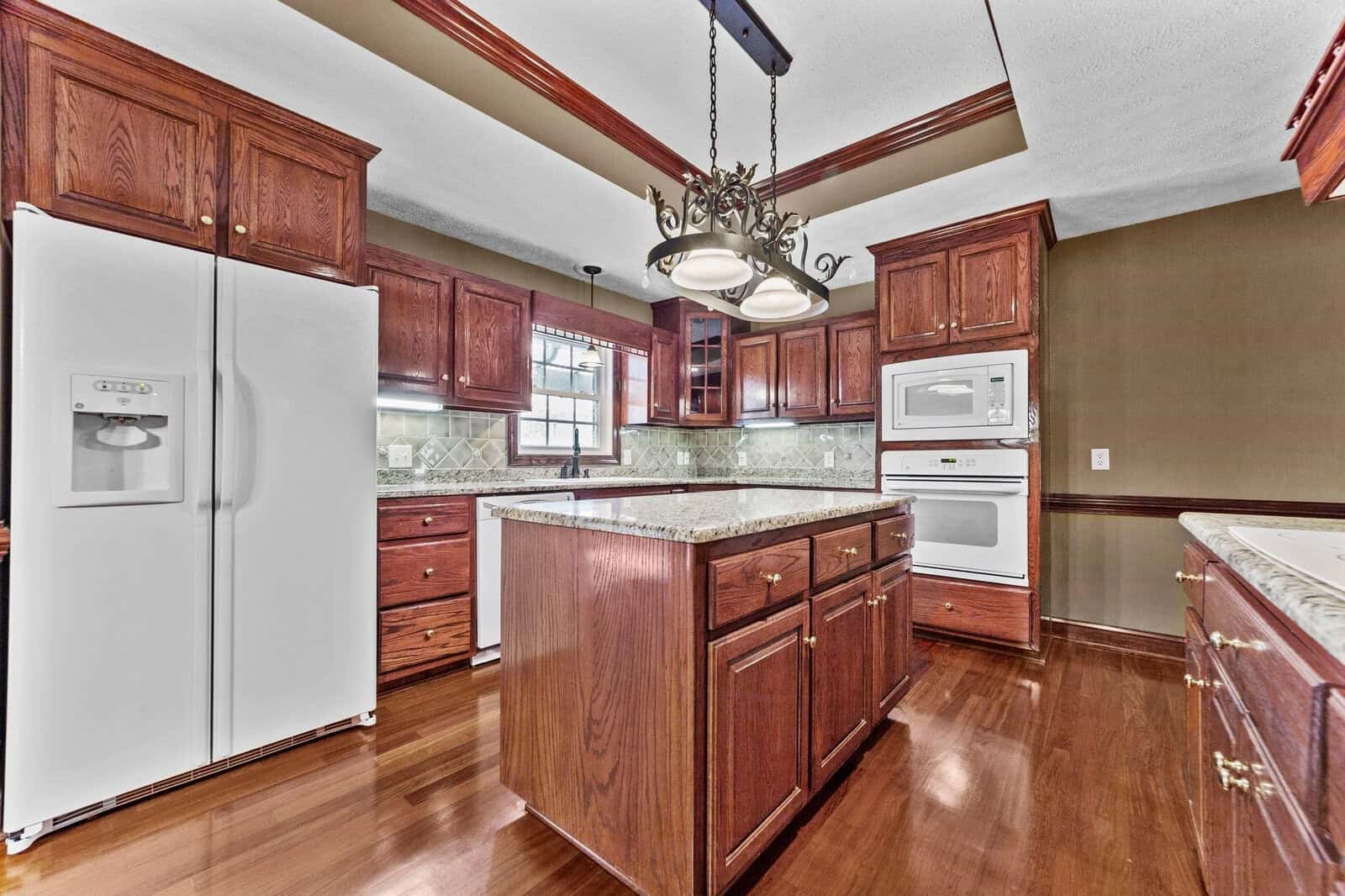 A spacious kitchen featuring cherry wood cabinets, granite countertops, white appliances, and a hanging chandelier above the island.