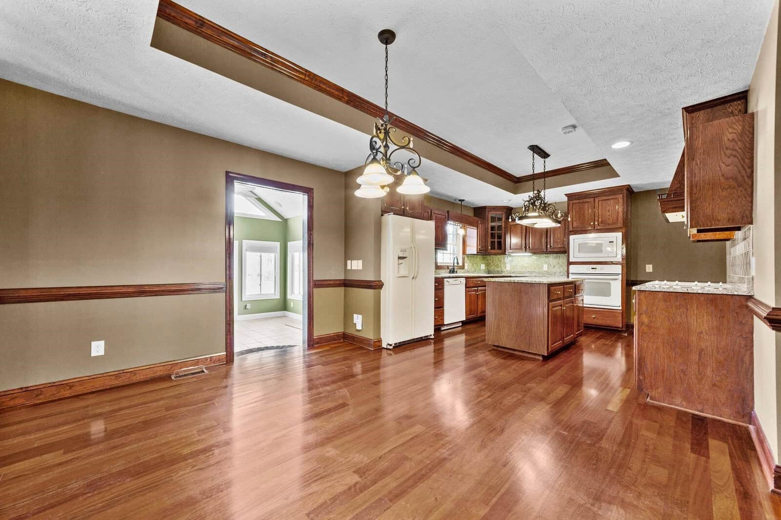 An empty dining room adjacent to a kitchen with wooden cabinets, granite countertops, and hardwood flooring.