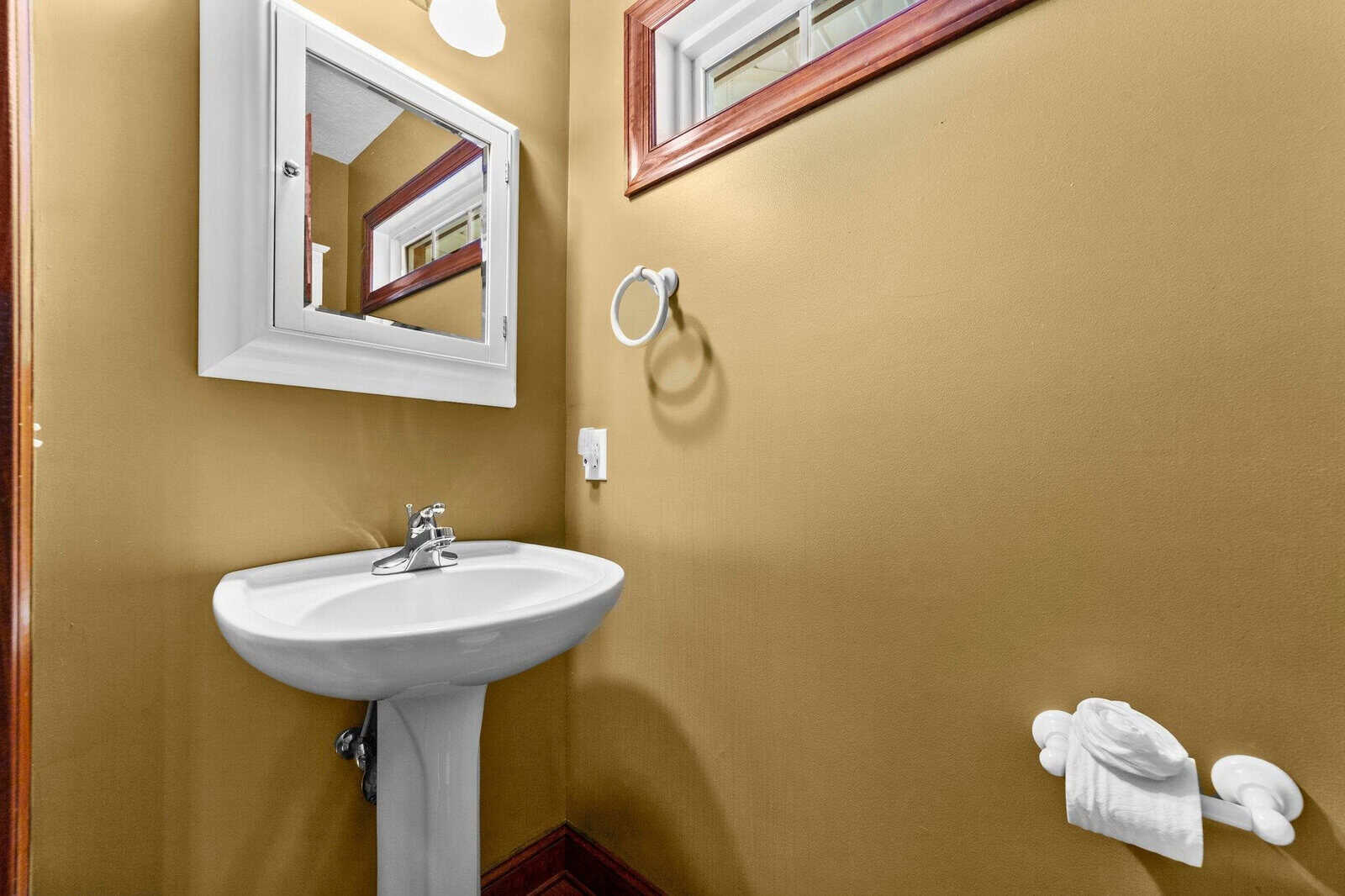 A small bathroom with mustard yellow walls featuring a pedestal sink, a mirror, and a towel holder with a white towel.