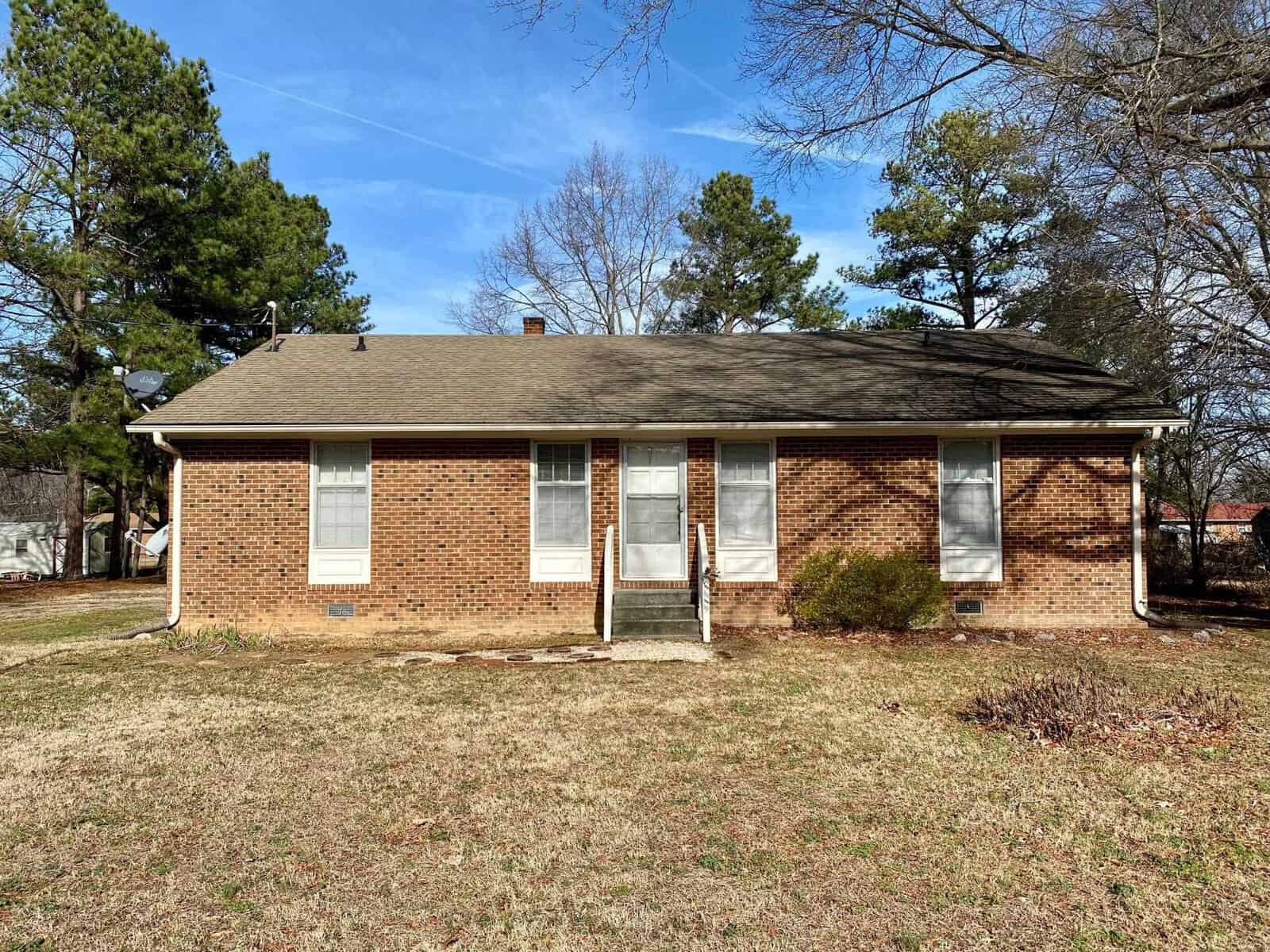 A single-story brick house with a shingled roof and a small front porch, set against a backdrop of leafless trees and a clear sky.