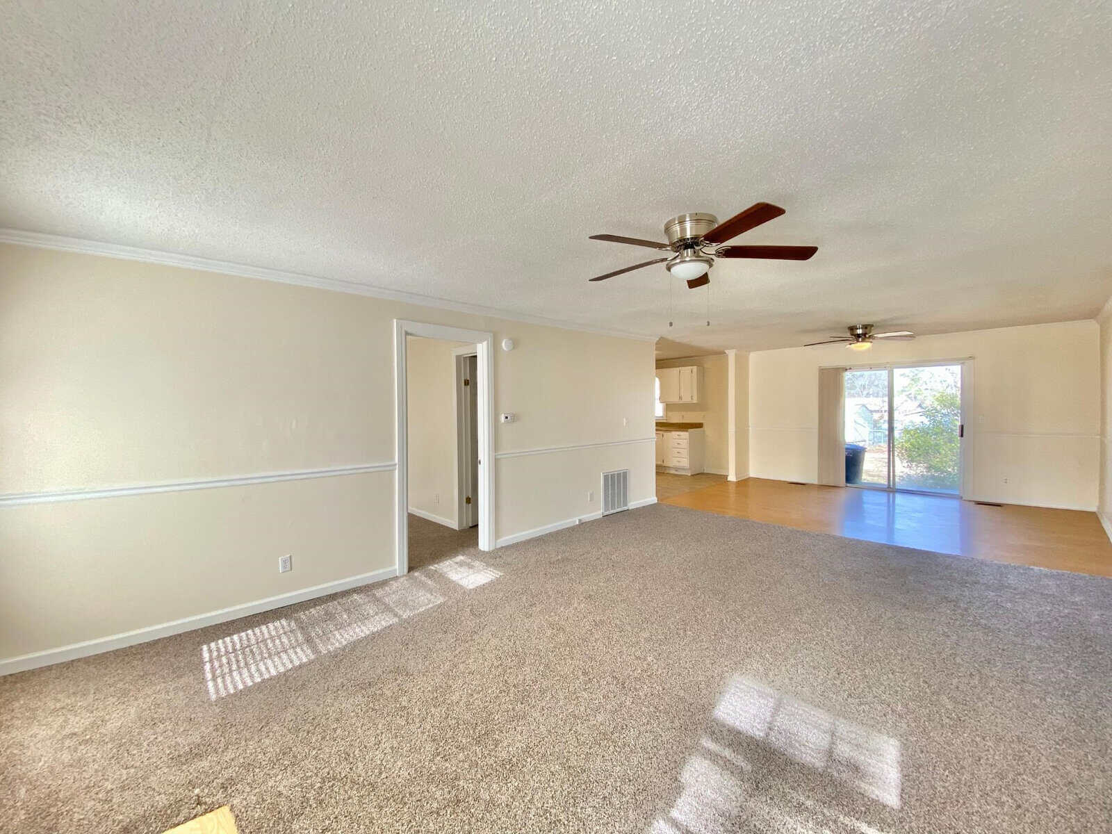 A spacious, empty room with beige walls and carpeted floor, featuring a ceiling fan and sunlight filtering through the windows.