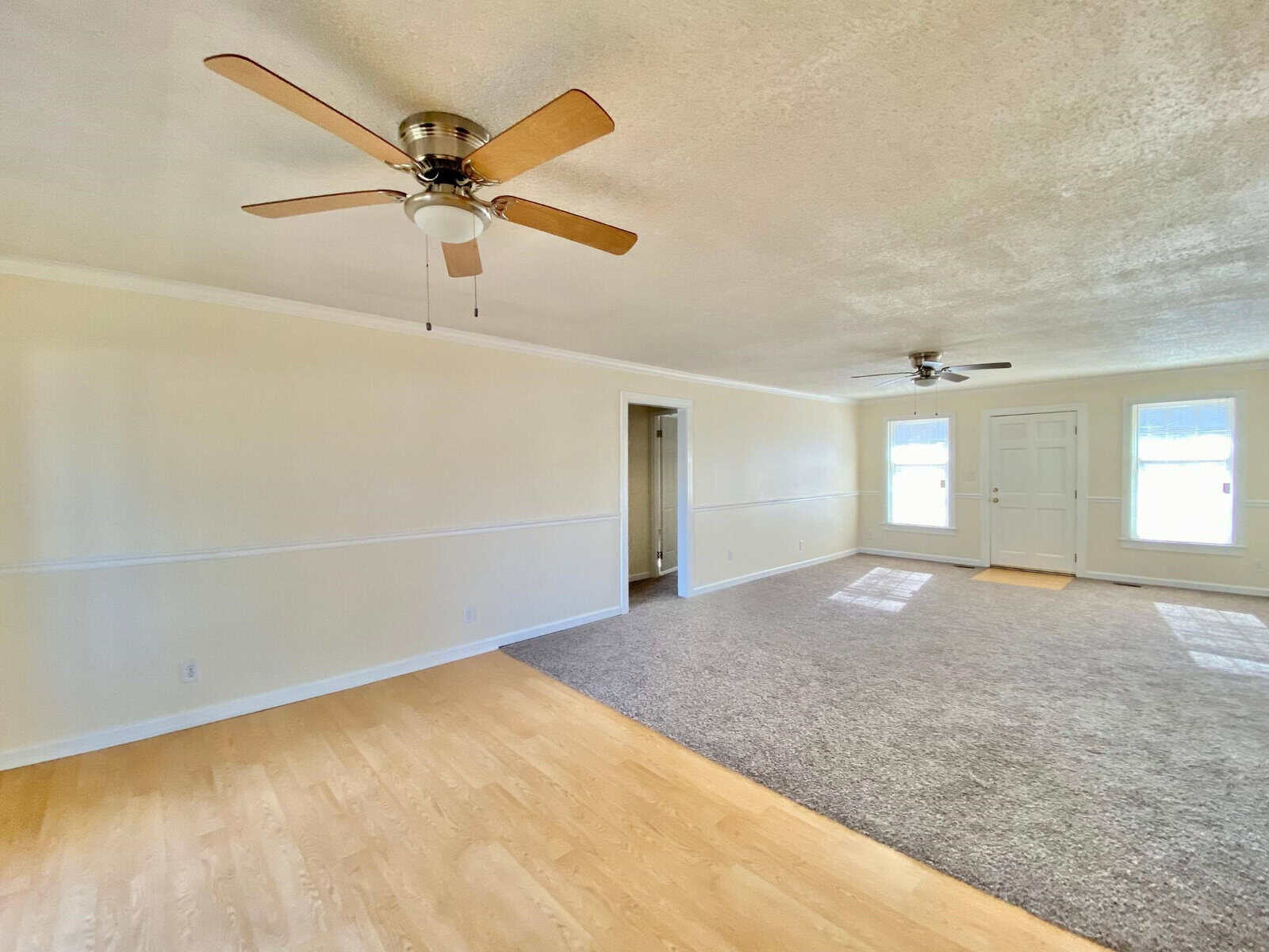 An empty room featuring a wooden floor in the foreground, carpeting in the back, a white chair rail dividing light-colored walls, and two ceiling fans.