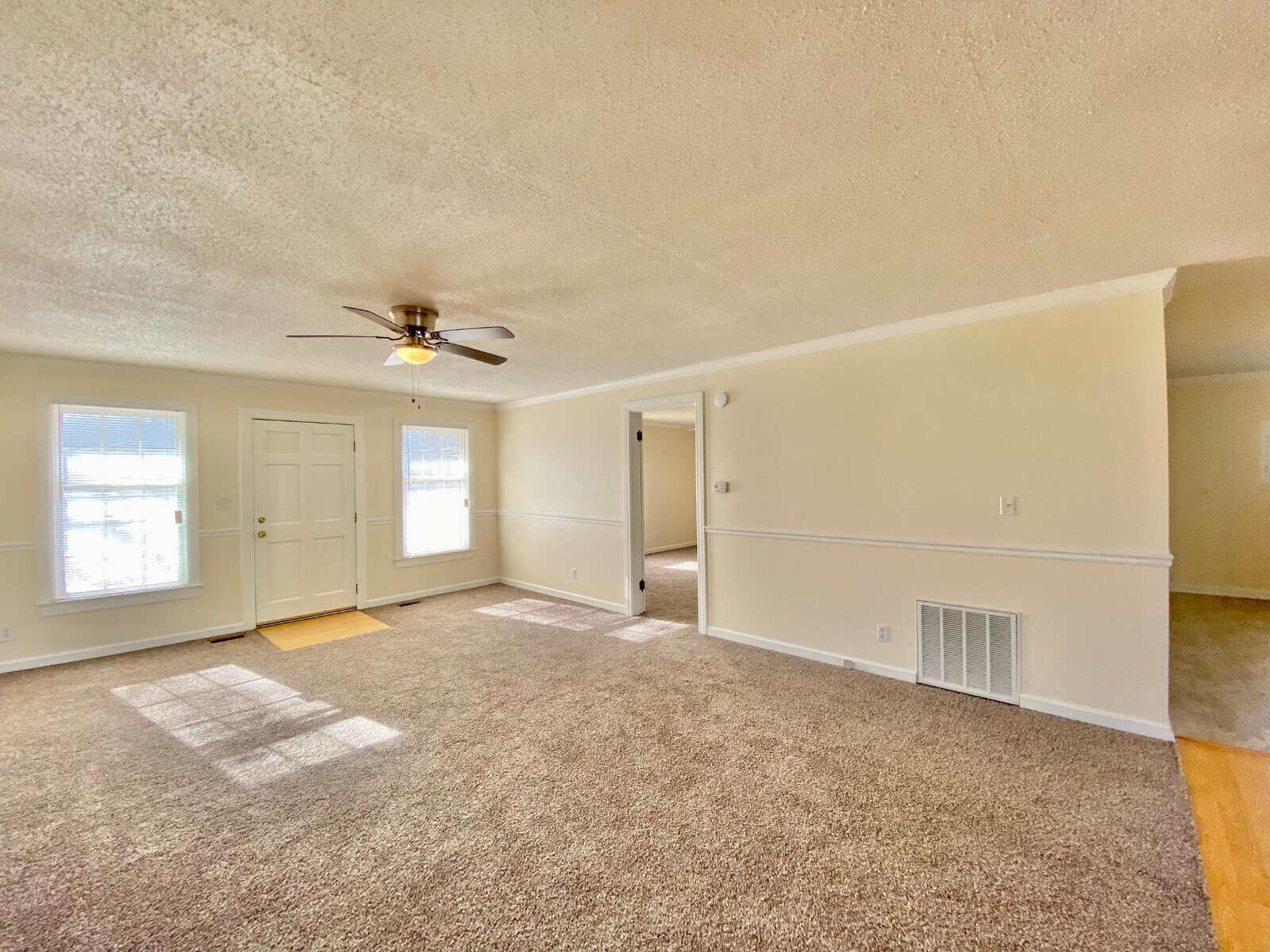 A spacious, empty room with beige carpeting, white walls, and a ceiling fan, featuring a front door with sidelight windows and an interior doorway.