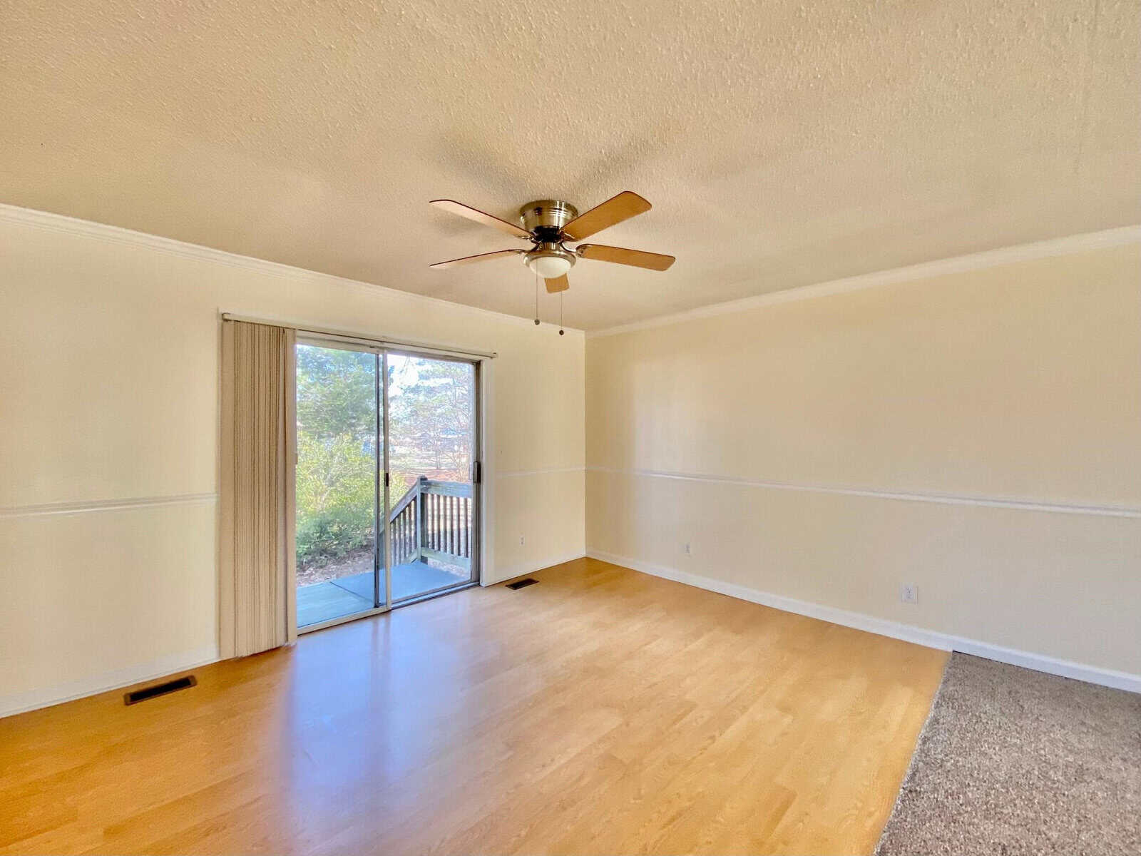An empty room with beige walls and hardwood flooring features a ceiling fan and a sliding glass door leading to an outdoor balcony.