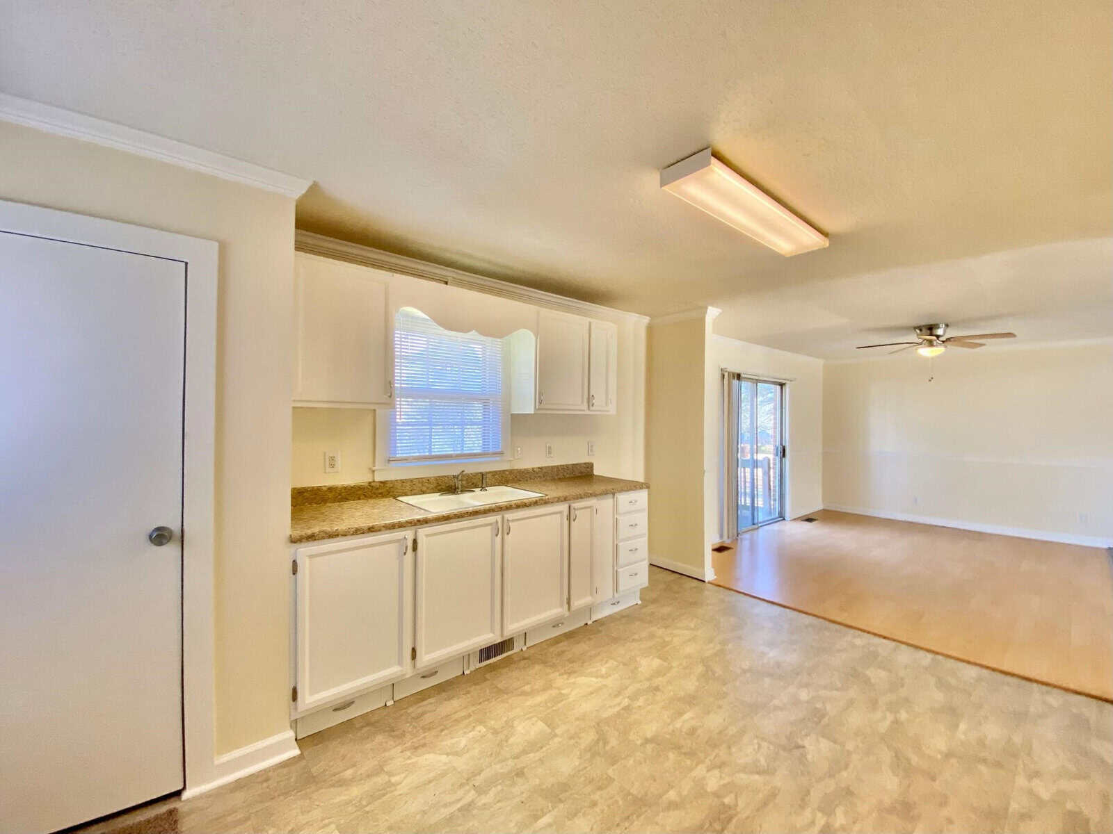 A bright, empty room featuring a kitchen area with white cabinetry and a living space with laminate flooring, illuminated by natural light from a window and a glass door.