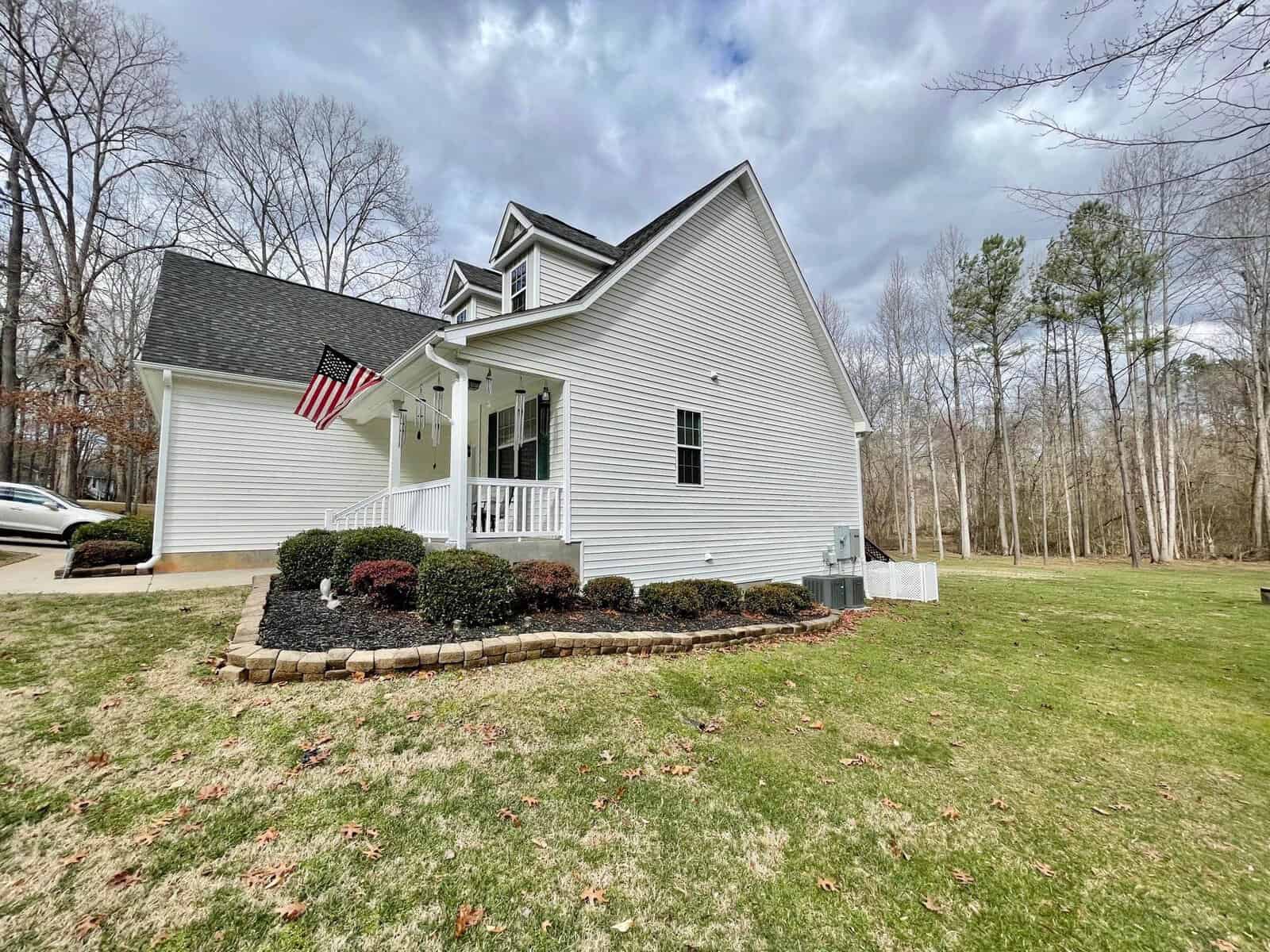 A white single-family house with a gabled roof featuring an american flag, situated in a wooded area with a well-maintained lawn and landscaping.