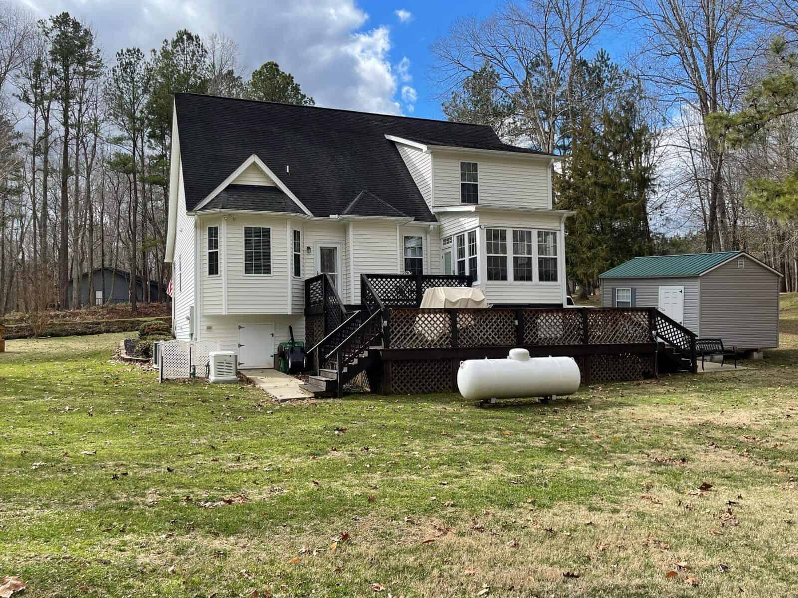 A two-story house with white siding and black shutters, featuring a large deck and a propane tank in the yard, set against a backdrop of trees and a partly cloudy sky.