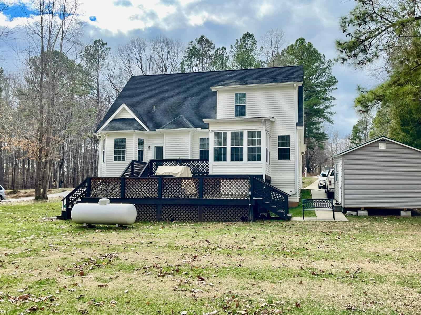 Two-story white house with black shutters and a front porch, featuring a propane tank on the side yard, adjacent to a detached garage or shed, surrounded by a lawn with scattered leaves and a tree-lined background.