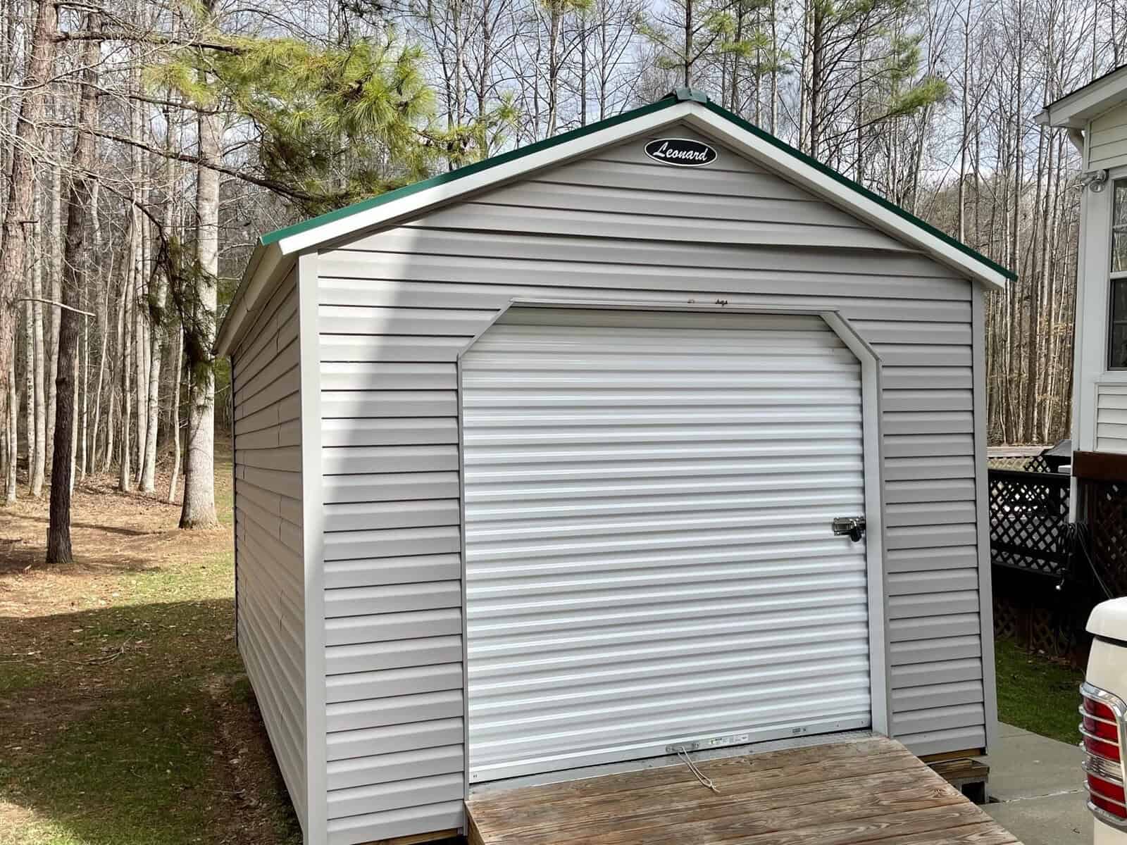 A vinyl-sided storage shed with a roll-up door situated in a wooded area.