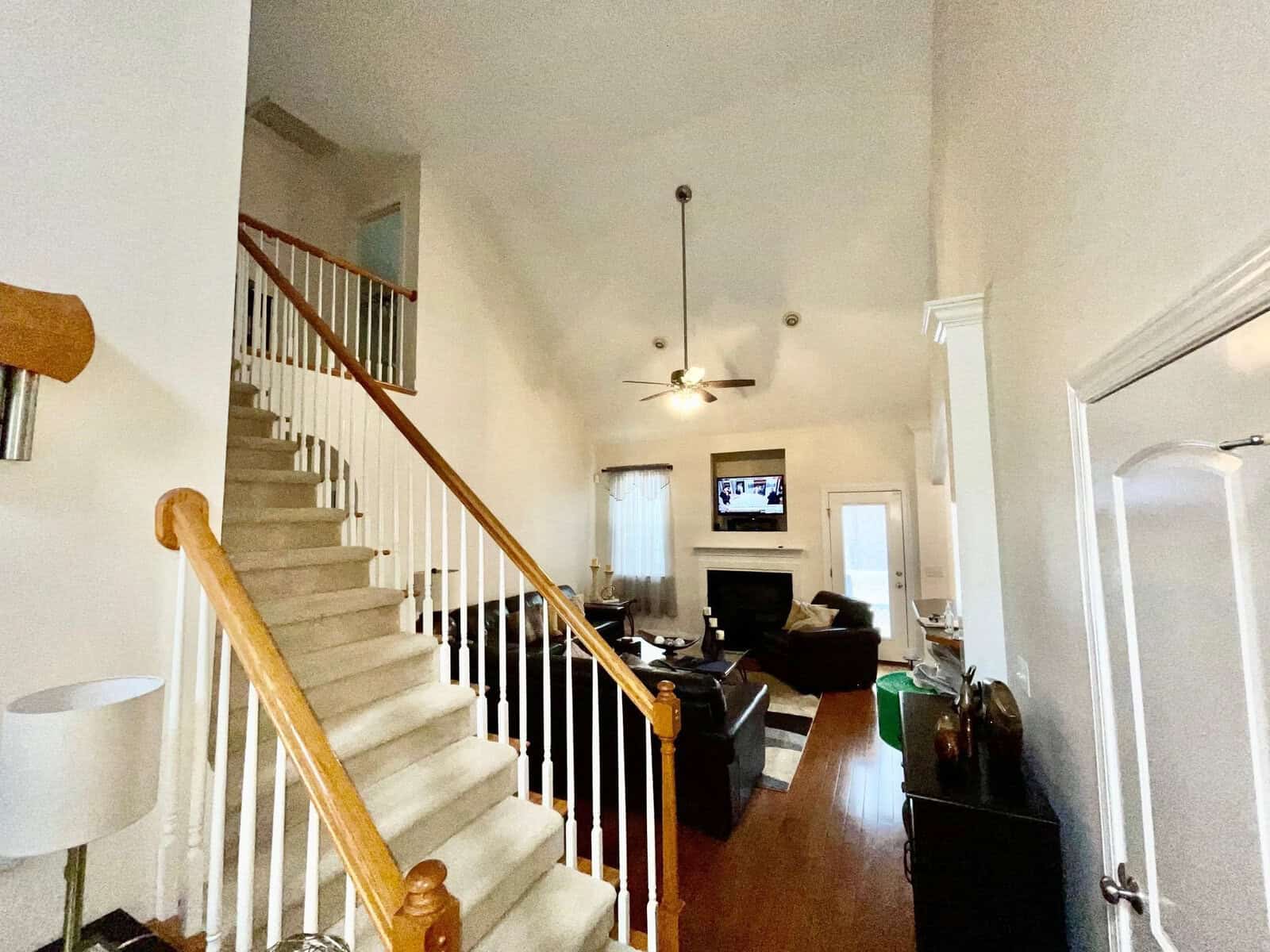 A well-lit interior of a house showing a staircase on the left leading to an upper level, with a living room area straight ahead featuring a ceiling fan, a television displaying an image, and various furnishings.