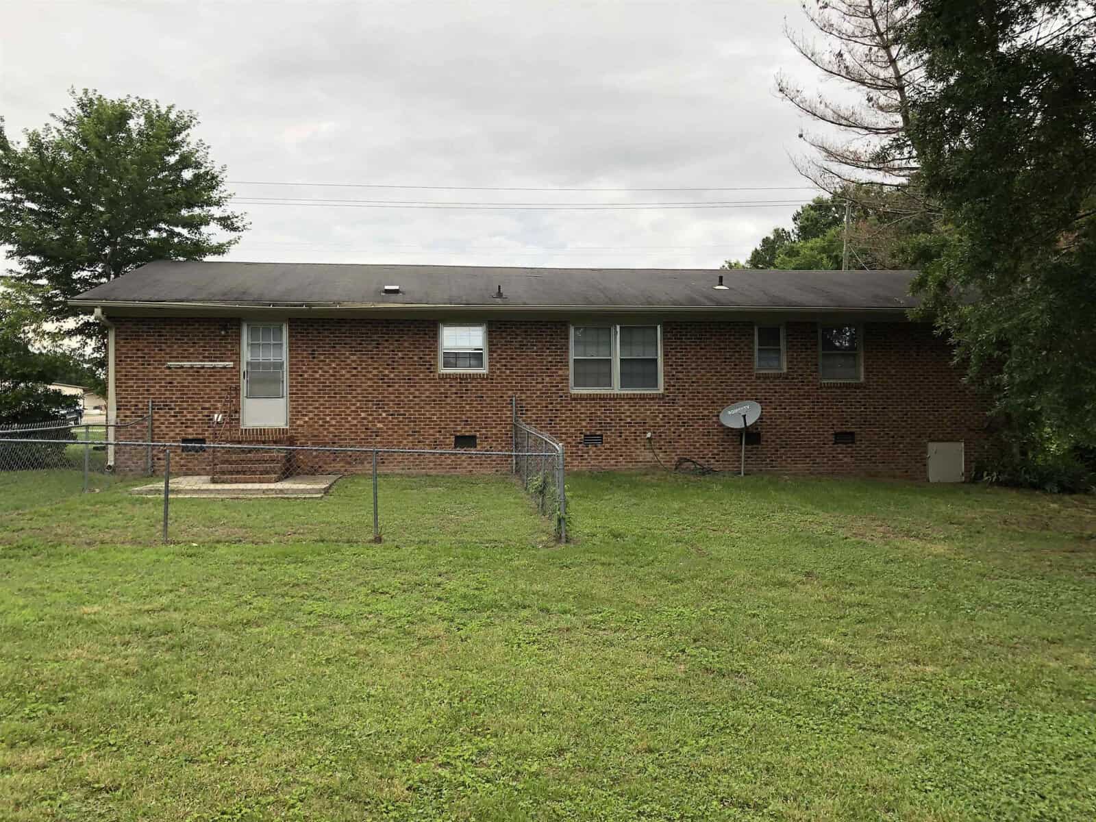 A single-story brick house with a dark roof, featuring a fenced front yard, a satellite dish, and a ramp leading to the front door, surrounded by green grass and trees under a cloudy sky.