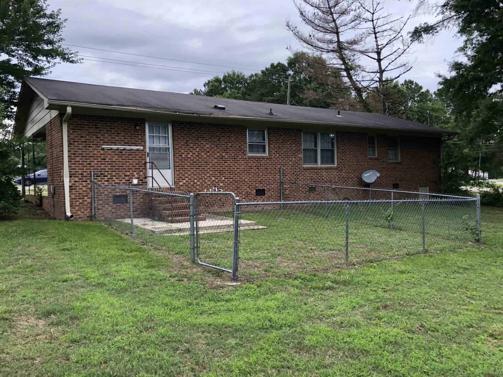 A single-story brick residential building with a fenced yard and a satellite dish on the side.