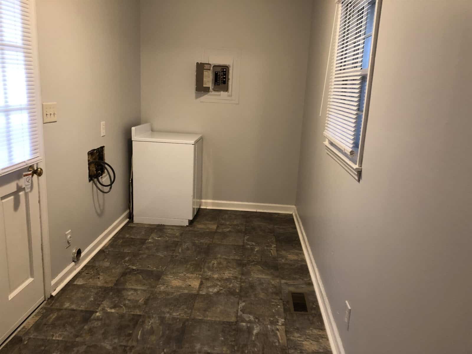 An empty room with beige walls, one window, and a patterned floor featuring a single white freezer and exposed utility connections on the left wall.