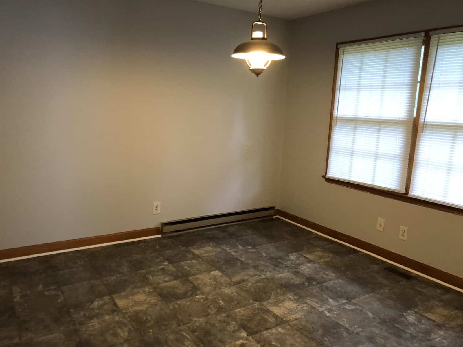 An empty room with gray walls, a hanging light fixture, and a brown patterned vinyl floor, featuring a window with closed blinds on one wall.