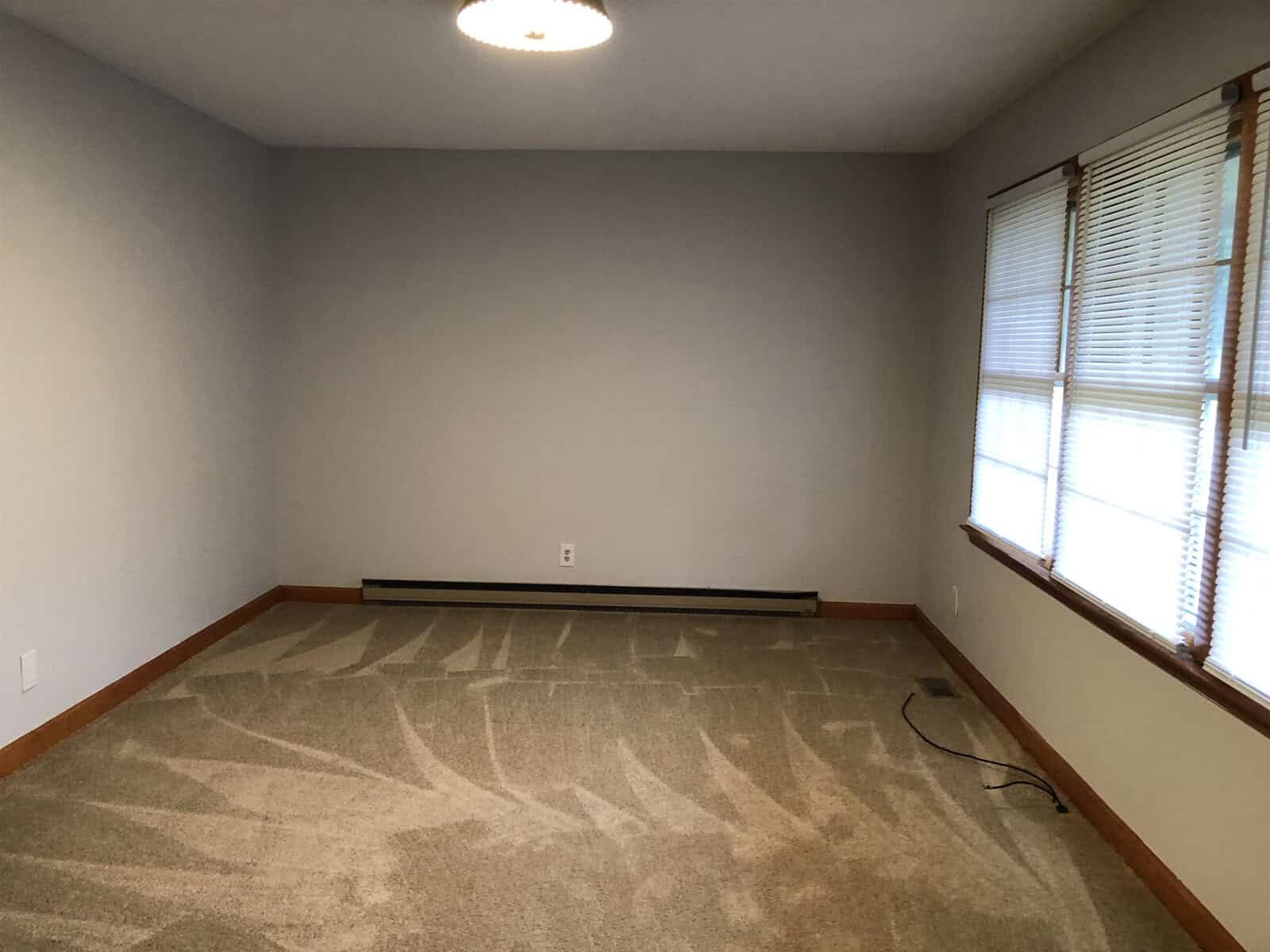 An empty room with beige walls, a patterned carpet, and one window with blinds.
