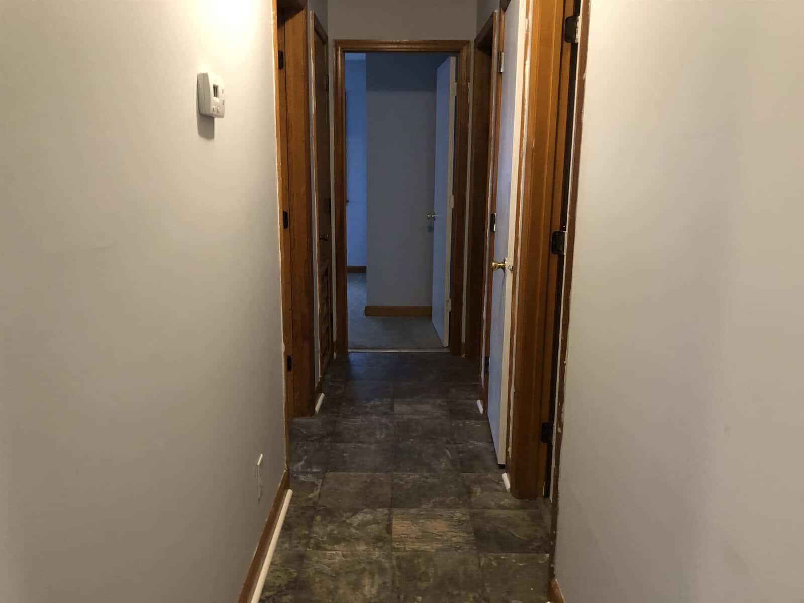 A dimly lit corridor with wooden doors on both sides leading to a room at the end with an open door.