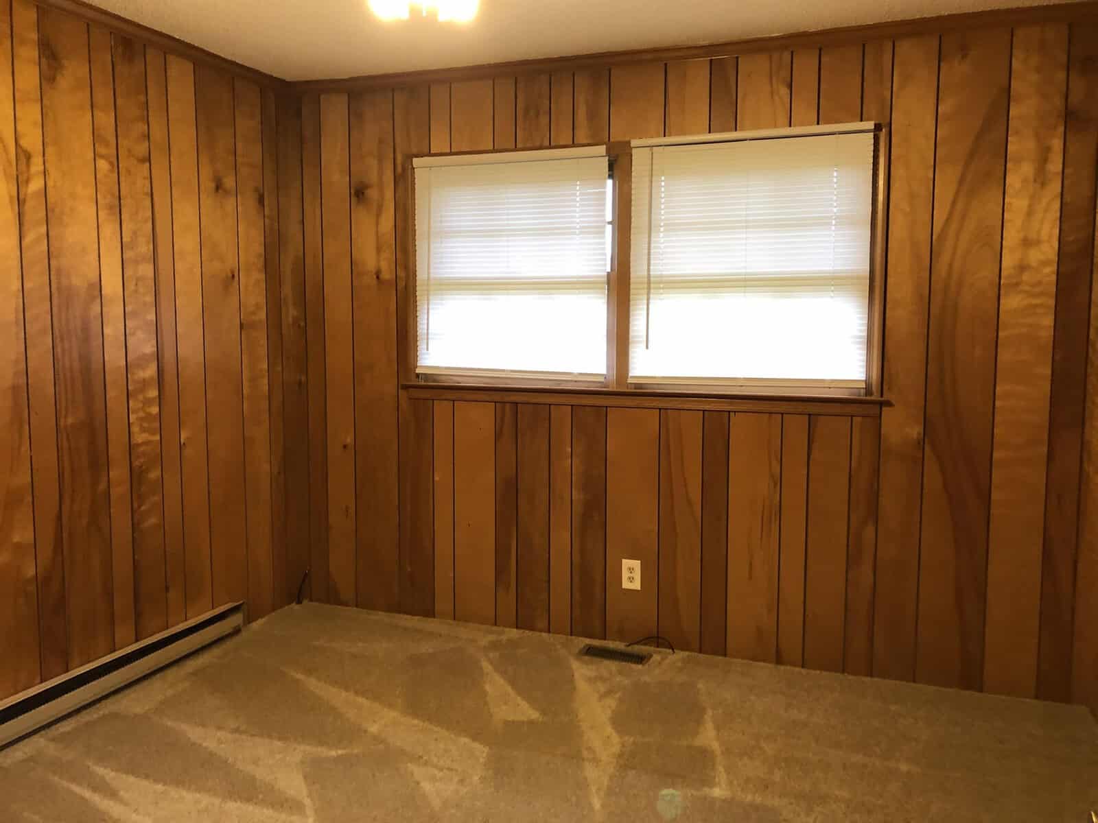 A room with wood paneling on the walls and two windows with horizontal blinds, featuring an unadorned beige carpeted floor.