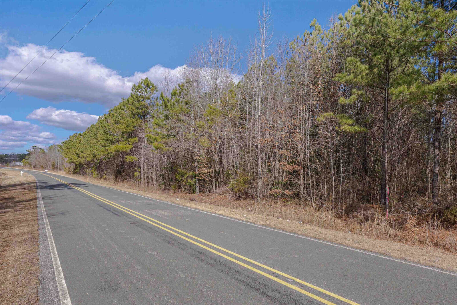 A two-lane road with a yellow dividing line running through a rural area with trees on either side under a partly cloudy sky.