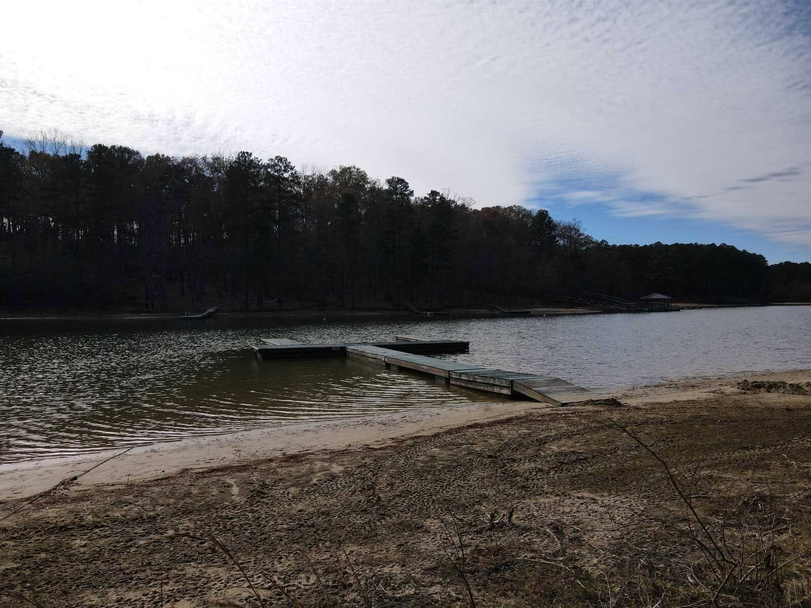 A calm lake with a small wooden dock extending into the water. The shoreline is sandy, and the background features a tree line of dense, bare trees under a partly cloudy sky.