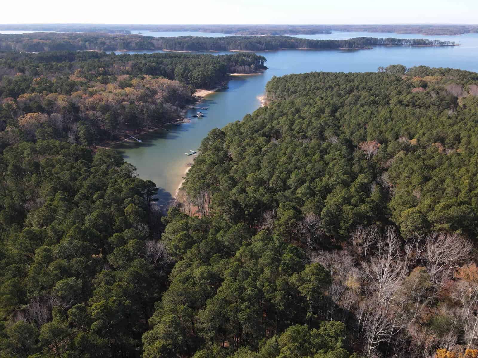 Aerial view of a large lake surrounded by lush green forests. A narrow inlet with calm water stretches into the forest, with a few boats visible near the shoreline. It's a clear day with a wide expanse of the lake in the background.