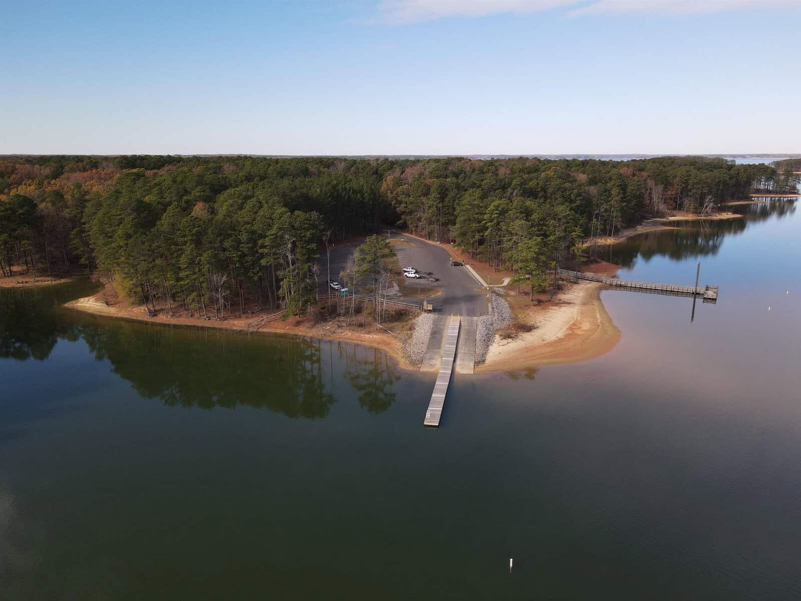 Aerial view of a serene lake with a small dock extending from a parking area into the water. Surrounded by trees, the area has a sandy shoreline on one side, and calm, clear water reflecting the sky on another. Sparse vehicles are visible in the lot.