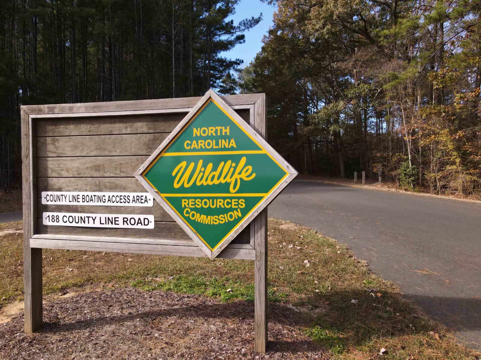 A wooden sign beside a paved road surrounded by trees reads "County Line Boating Access Area, 188 County Line Road." Another sign, a diamond shape in green and yellow, reads "North Carolina Wildlife Resources Commission.