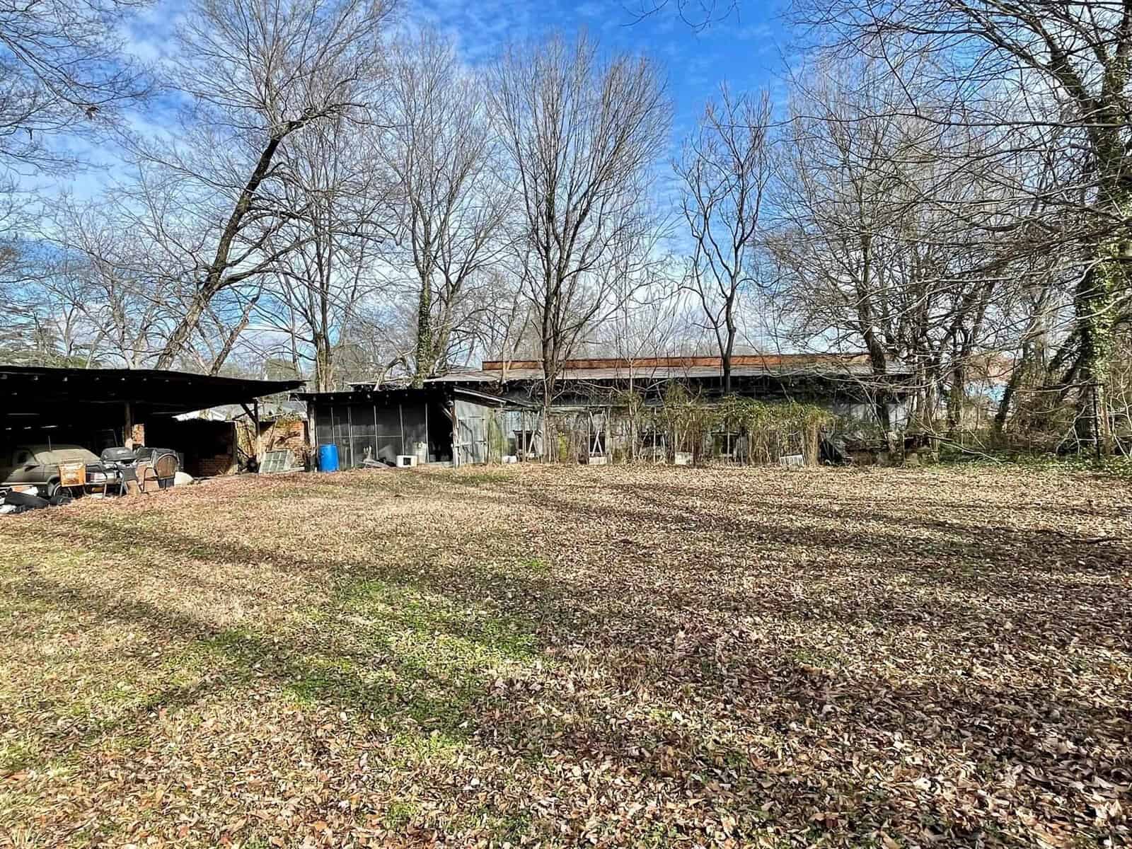 An open field with dry grass and scattered leaf litter, leading to a covered area with various items and structures, surrounded by leafless trees under a clear sky.