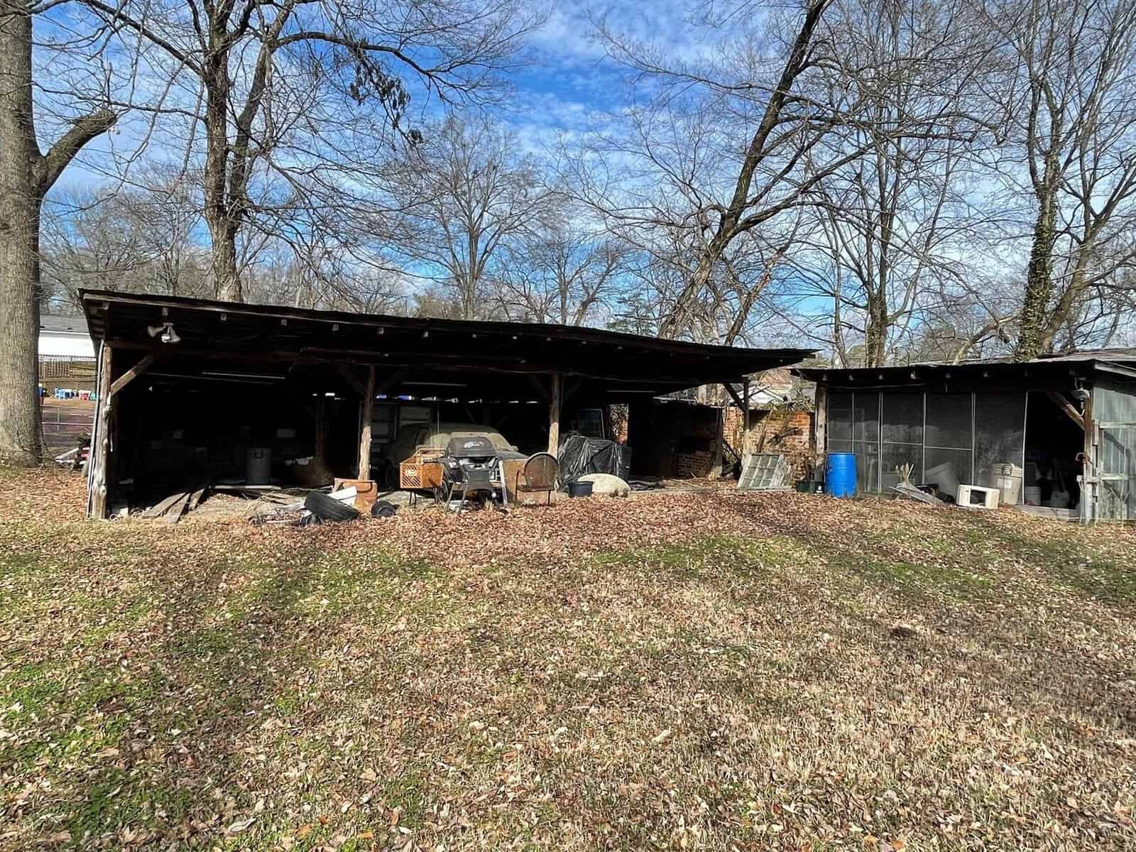 A rustic outdoor area with a covered carport containing various items, including a grill and storage boxes, adjacent to a small shed, surrounded by leaf-strewn grass and trees.