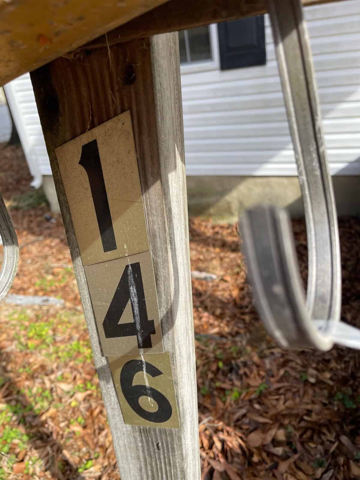 Reflective house numbers "146" are affixed to a weathered wooden post with a blurred background featuring a residential building.