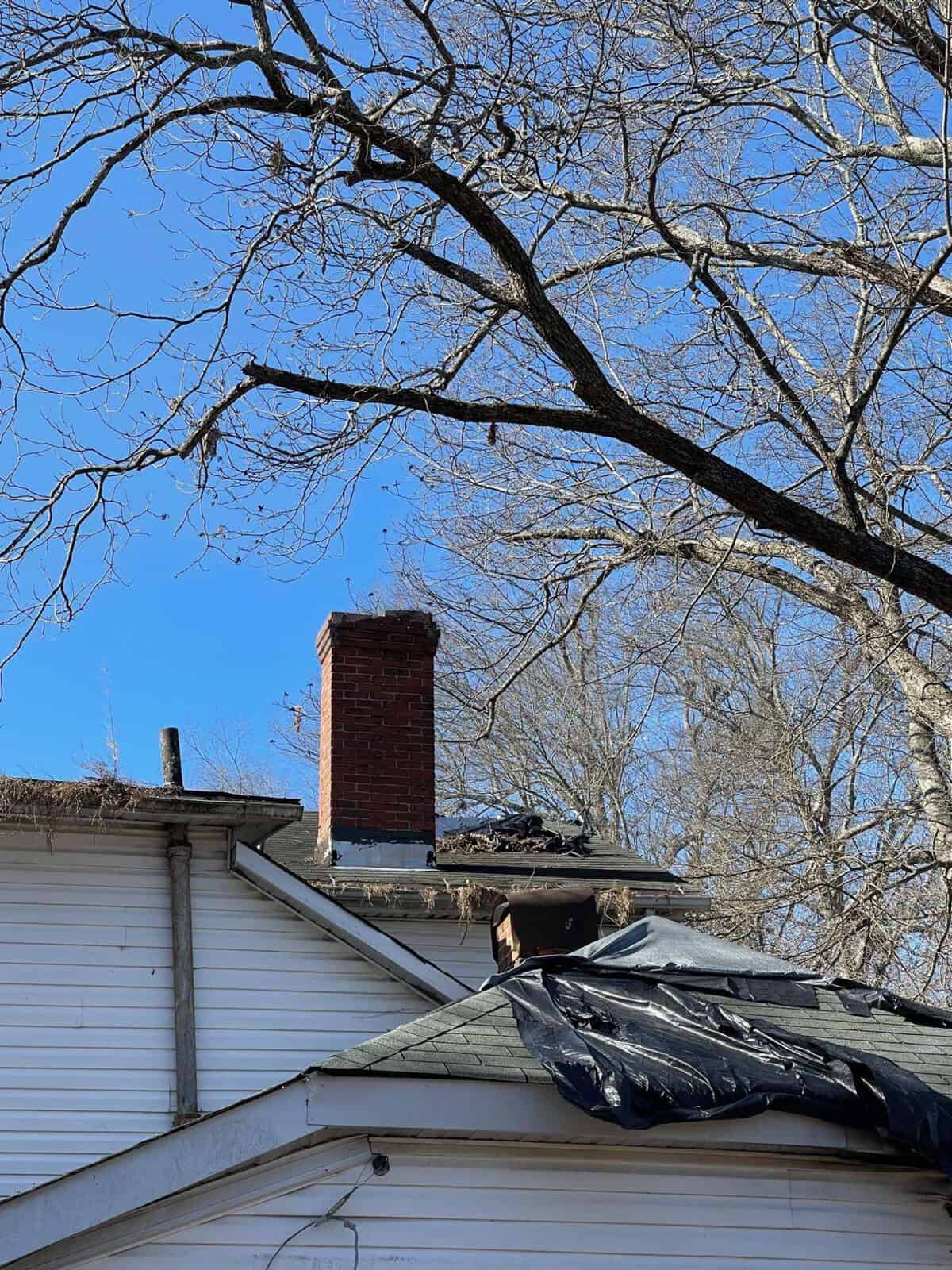 A red brick chimney stands atop a house with white siding; a tarp is partially draped over the roof, with leafless branches against a clear blue sky overhead.