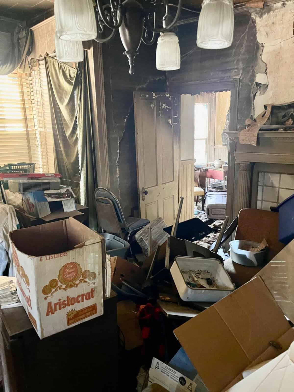 A cluttered room with various items strewn about, including boxes, furniture, and a disheveled chandelier, showing signs of neglect and disrepair.