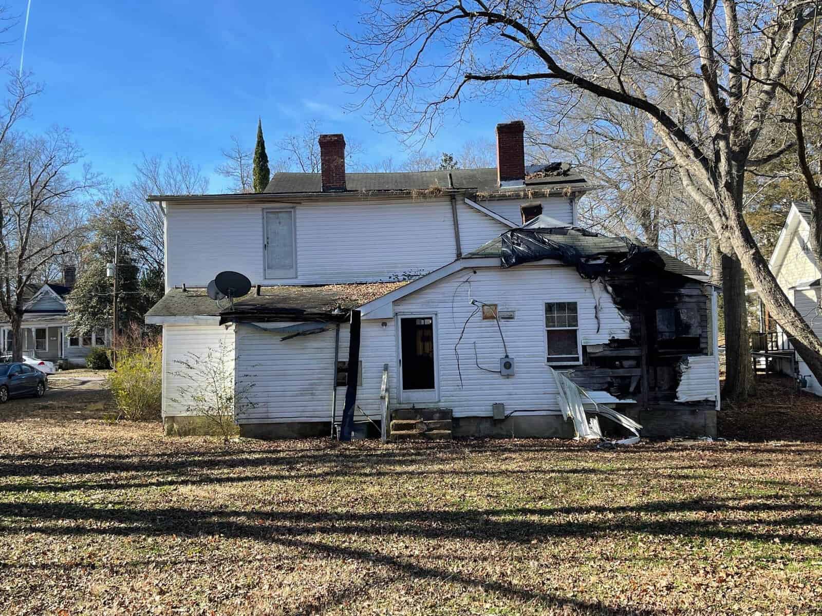 A white, two-story house with significant fire damage and debris on the back. The yard is leaf-covered, and nearby houses are visible under a blue sky with bare trees.