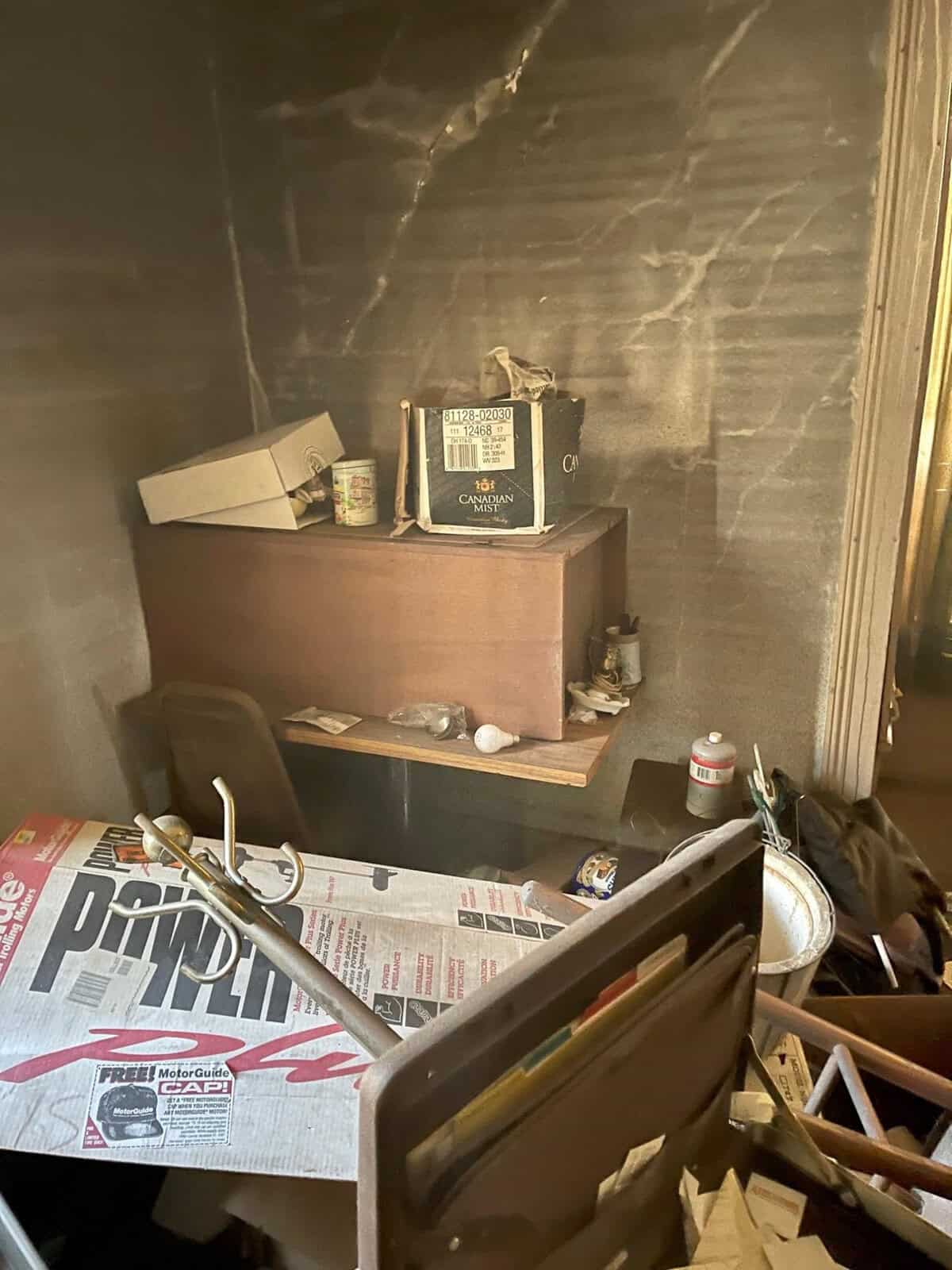 A small, cluttered room with smoke-stained walls, an overturned chair, scattered papers, and boxes on a desk. The scene appears disordered and affected by possible fire damage.