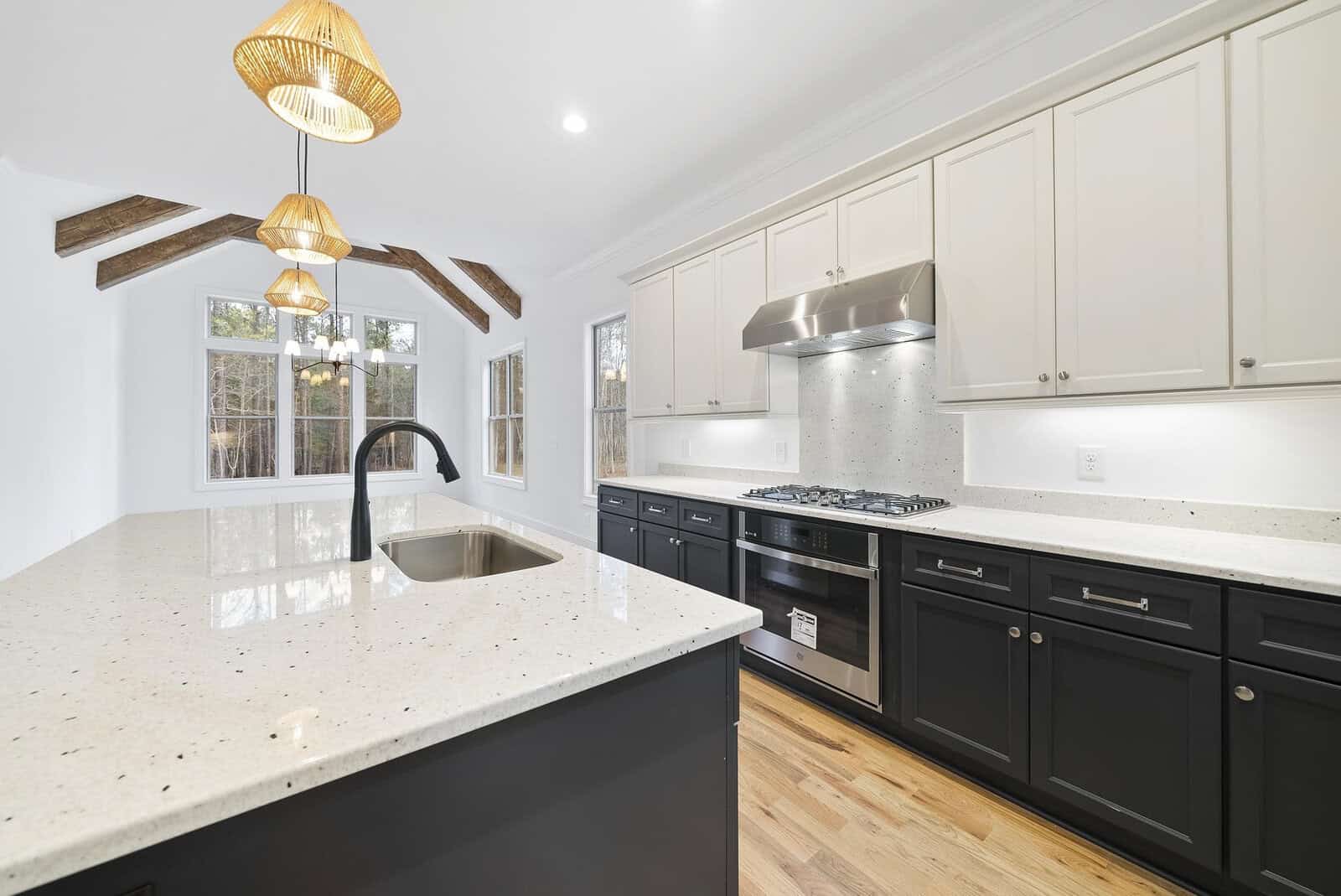 A modern kitchen featuring white cabinets, a stainless steel oven with a matching hood, and a large island with a sink. The ceiling has wooden beams and pendant lights, and large windows let in natural light.