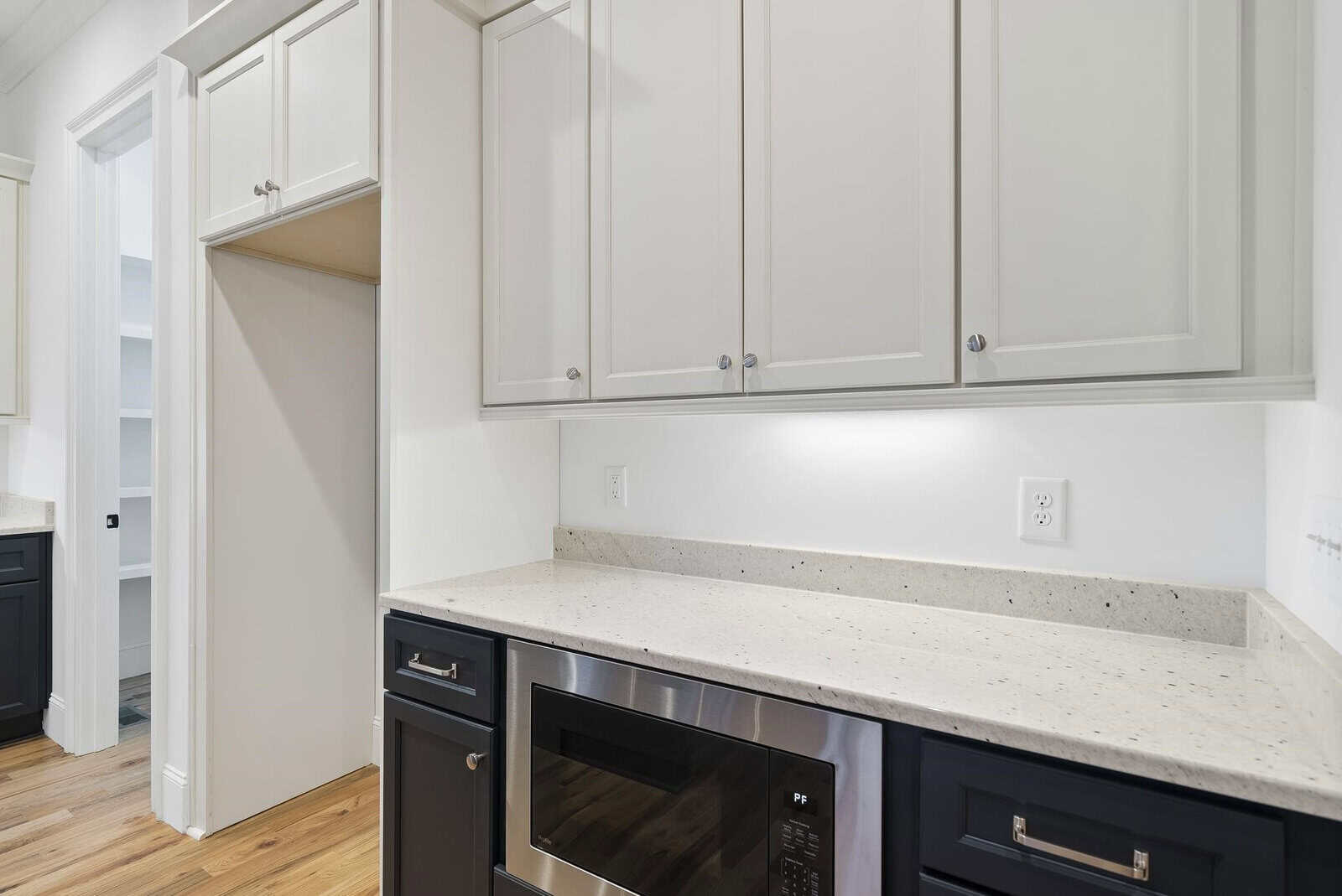 A modern kitchen features white upper cabinets, black lower cabinets, and a speckled countertop. A stainless steel microwave is built into the lower cabinets, and a pantry is partially visible through an open door.
