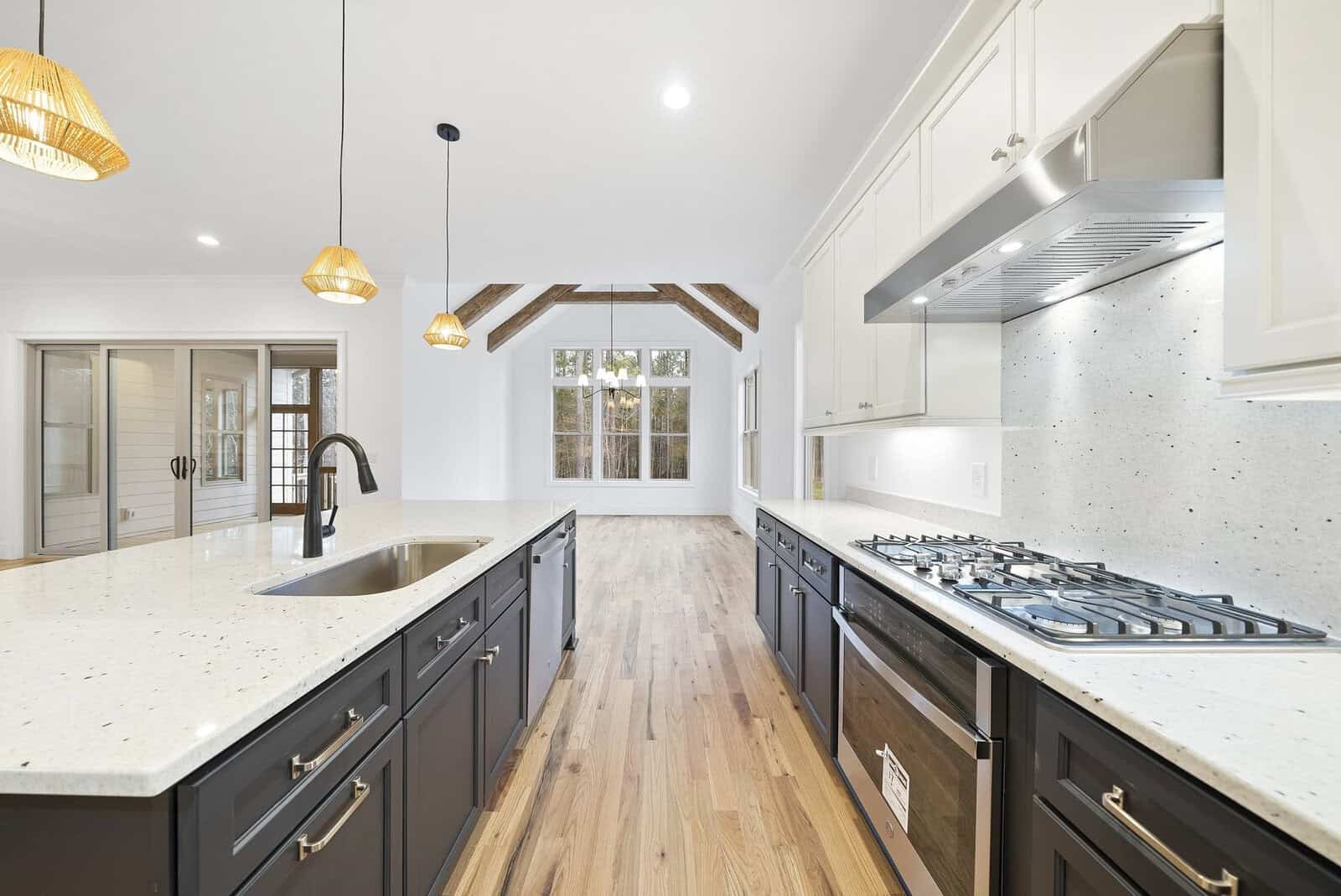 Spacious modern kitchen with hardwood floors, featuring a large island with a sink and dark cabinets. A gas stovetop is on the right, beneath stainless steel range hood. Pendant lights hang above, and windows offer natural light.