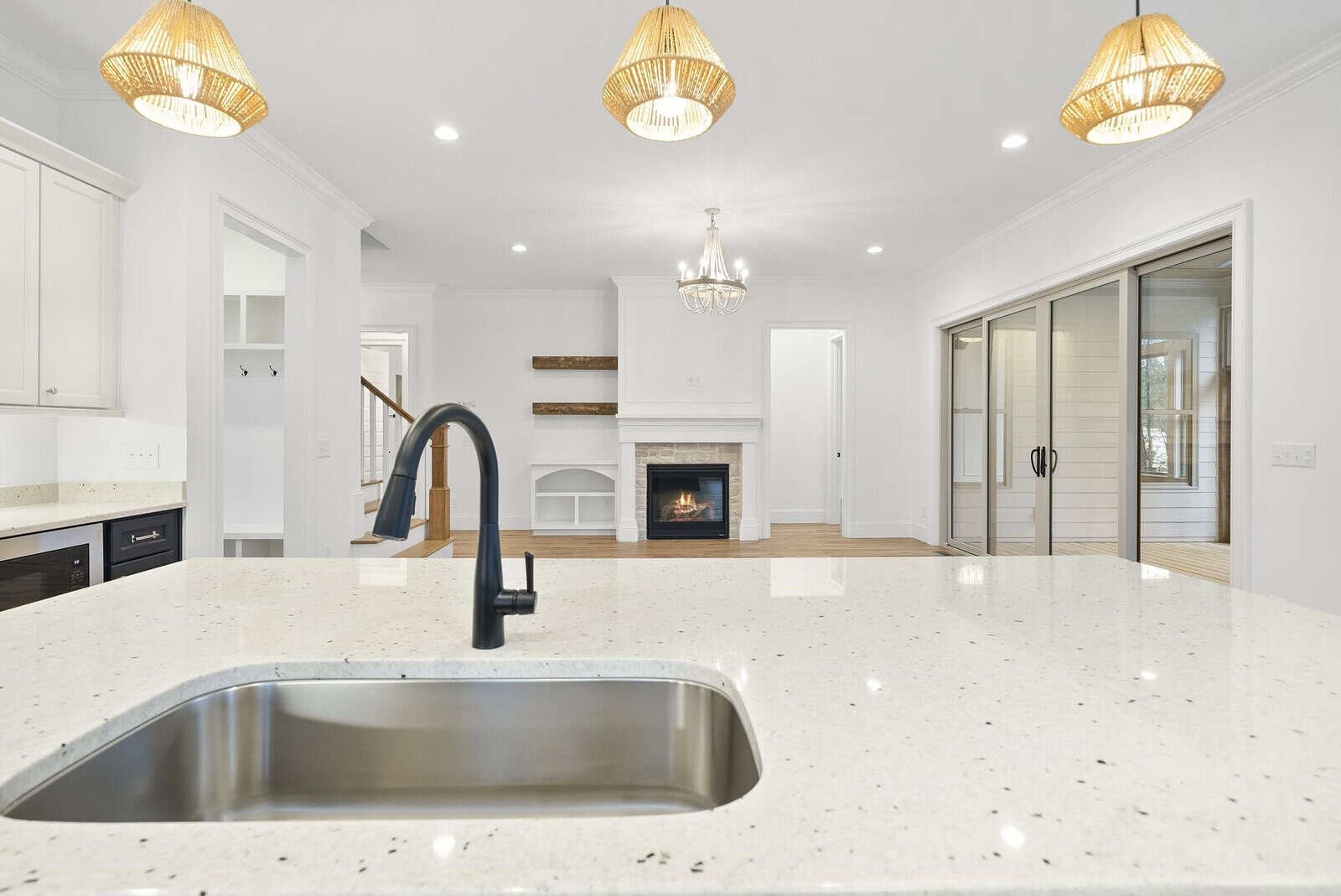 Modern kitchen with a large white countertop island featuring a black sink and faucet. Three pendant lights hang above, and a lit fireplace is visible in the background. Sliding glass doors on the right open to another room.