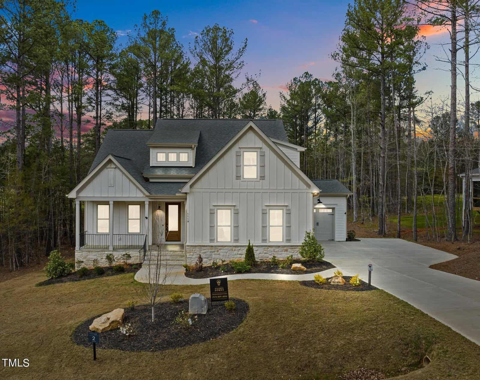 A modern two-story house at 1119 Springdale Drive in Wake Forest boasts a light exterior, surrounded by a manicured lawn and trees. The sunset sky's pink and blue hues add warmth to the serene landscape, while a driveway leads to the side garage.