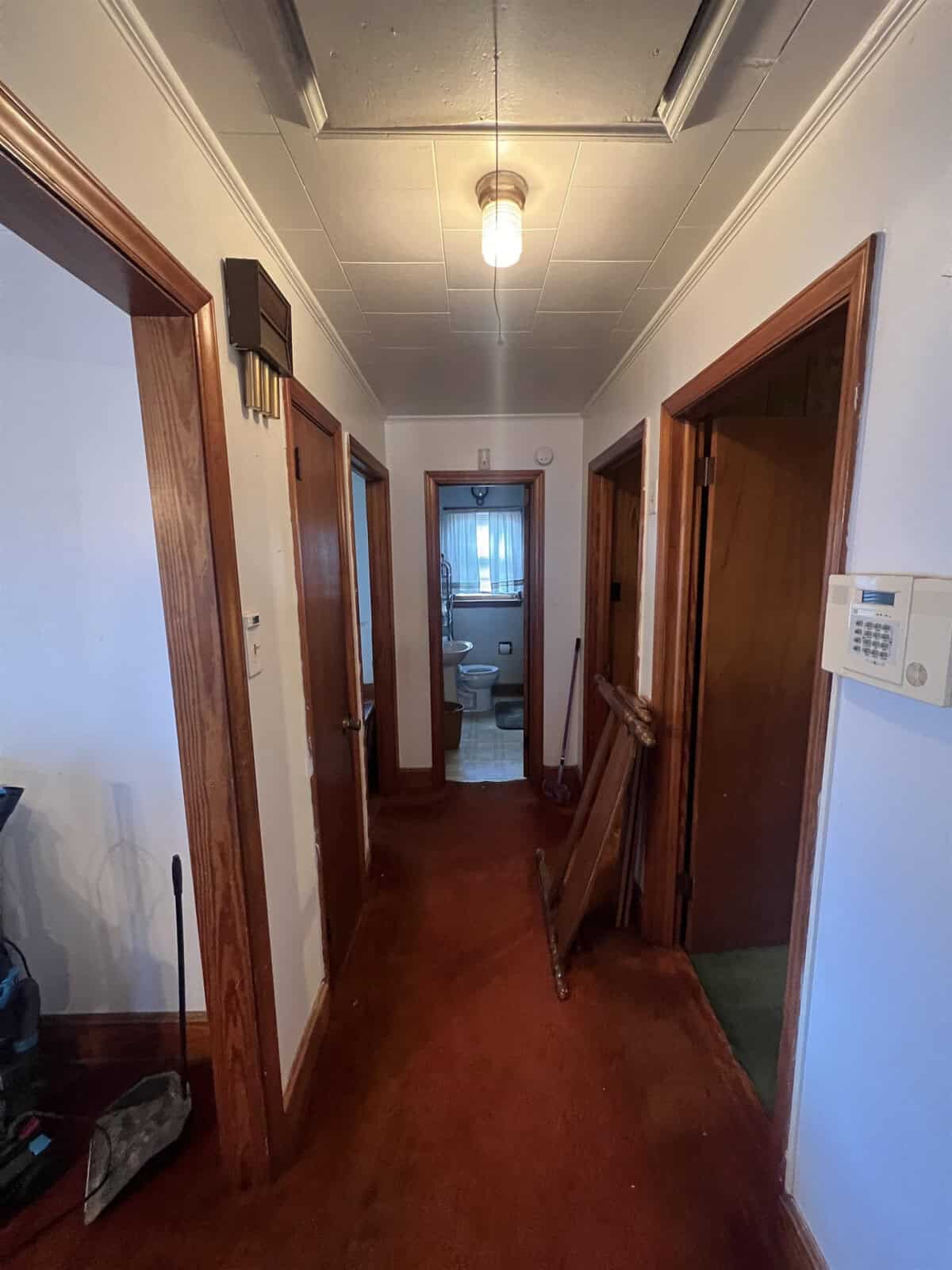 A narrow hallway with red carpeting, leading to a bathroom at the end, with doors on either side and a ceiling light fixture. there is a wooden cane leaning against the wall to the right.